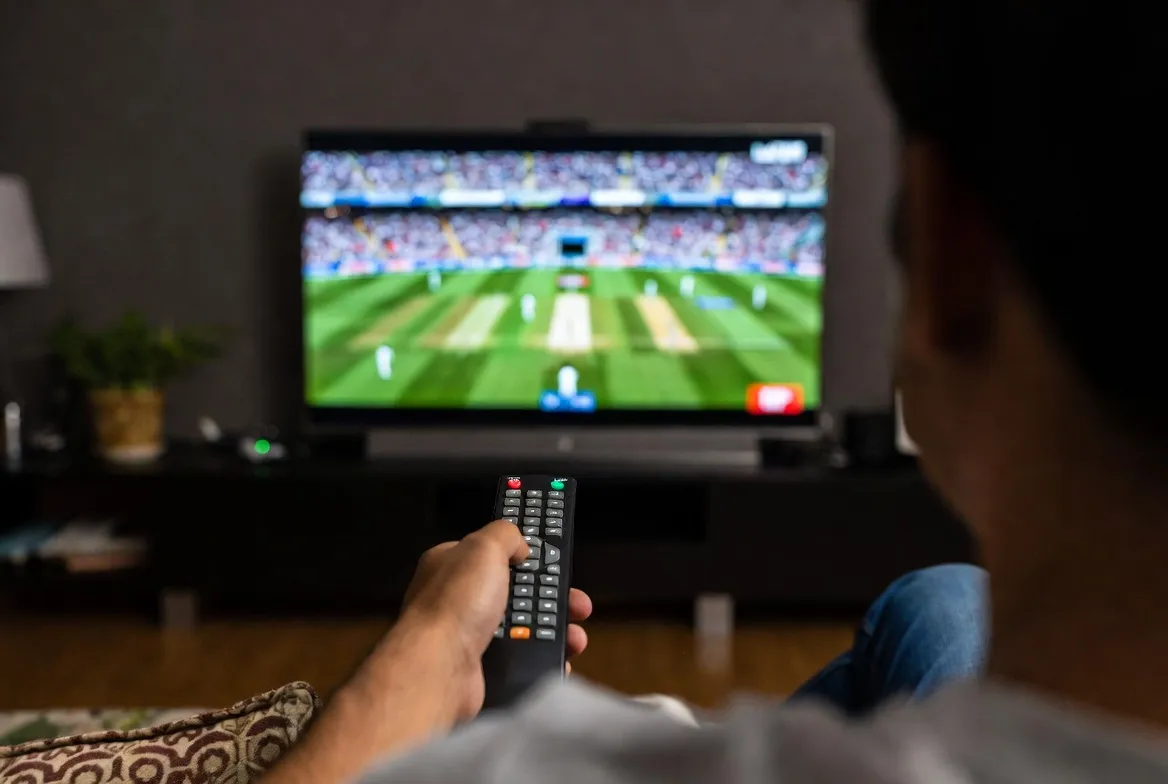 Viewer watching a cricket match live on TV with IPTV remote in hand
